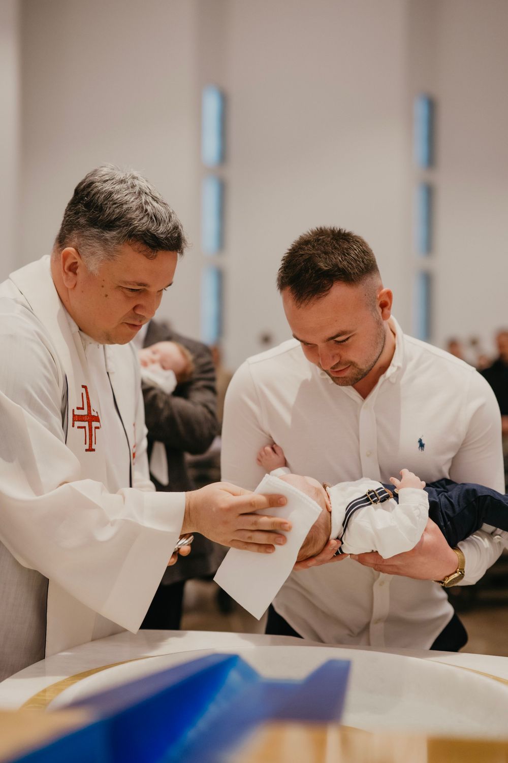 A priest and parent participate in a baptism ceremony while holding a small infant.