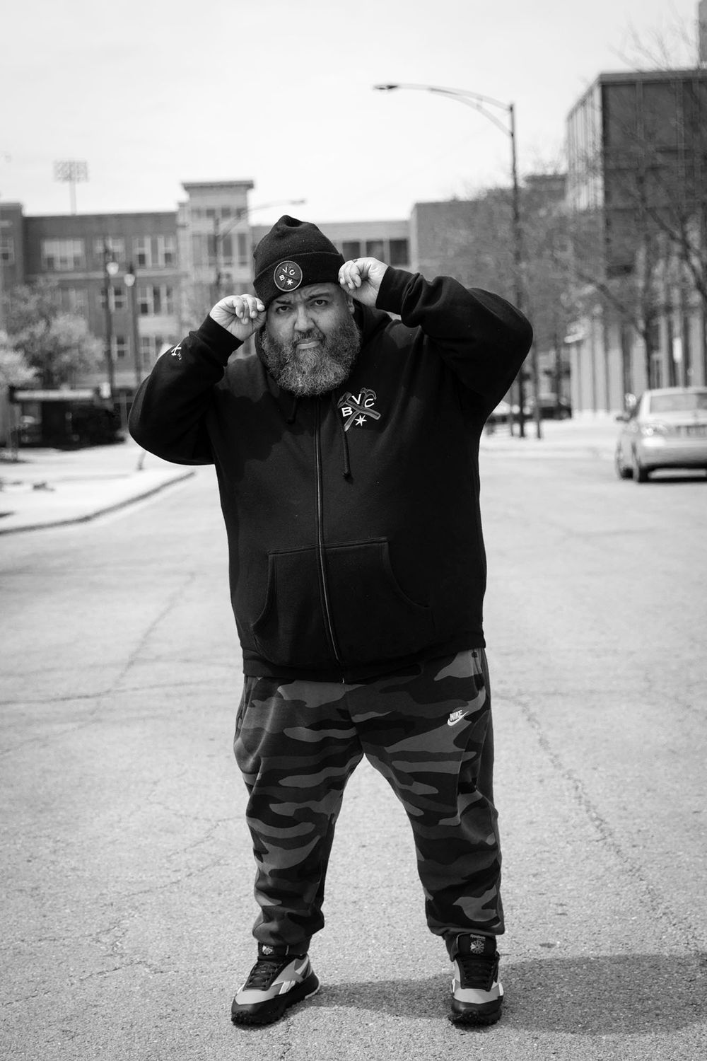 Street photography of person in camo pants and hoodie posing against urban backdrop.