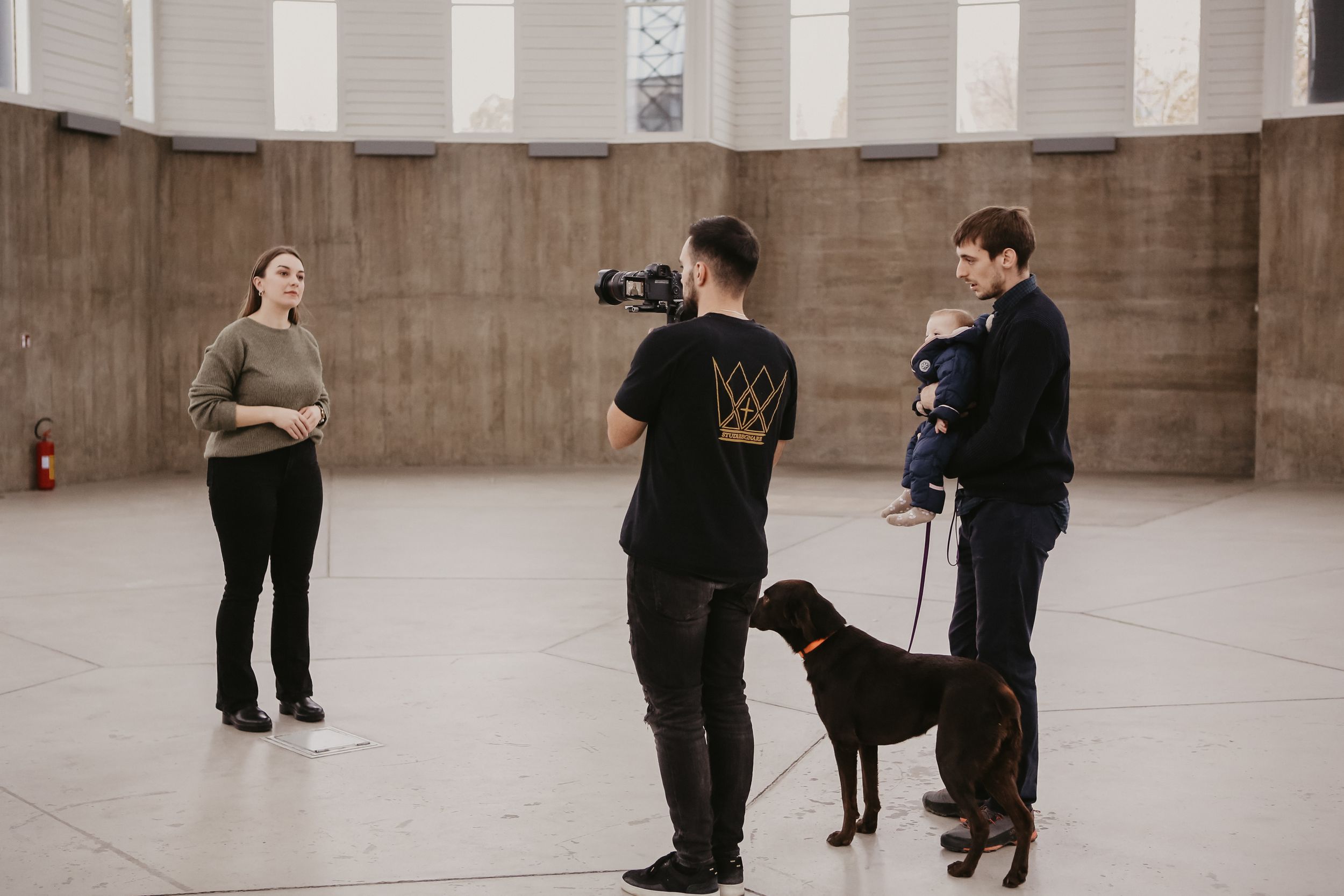 Two people with camera equipment and a black dog in a spacious indoor venue.