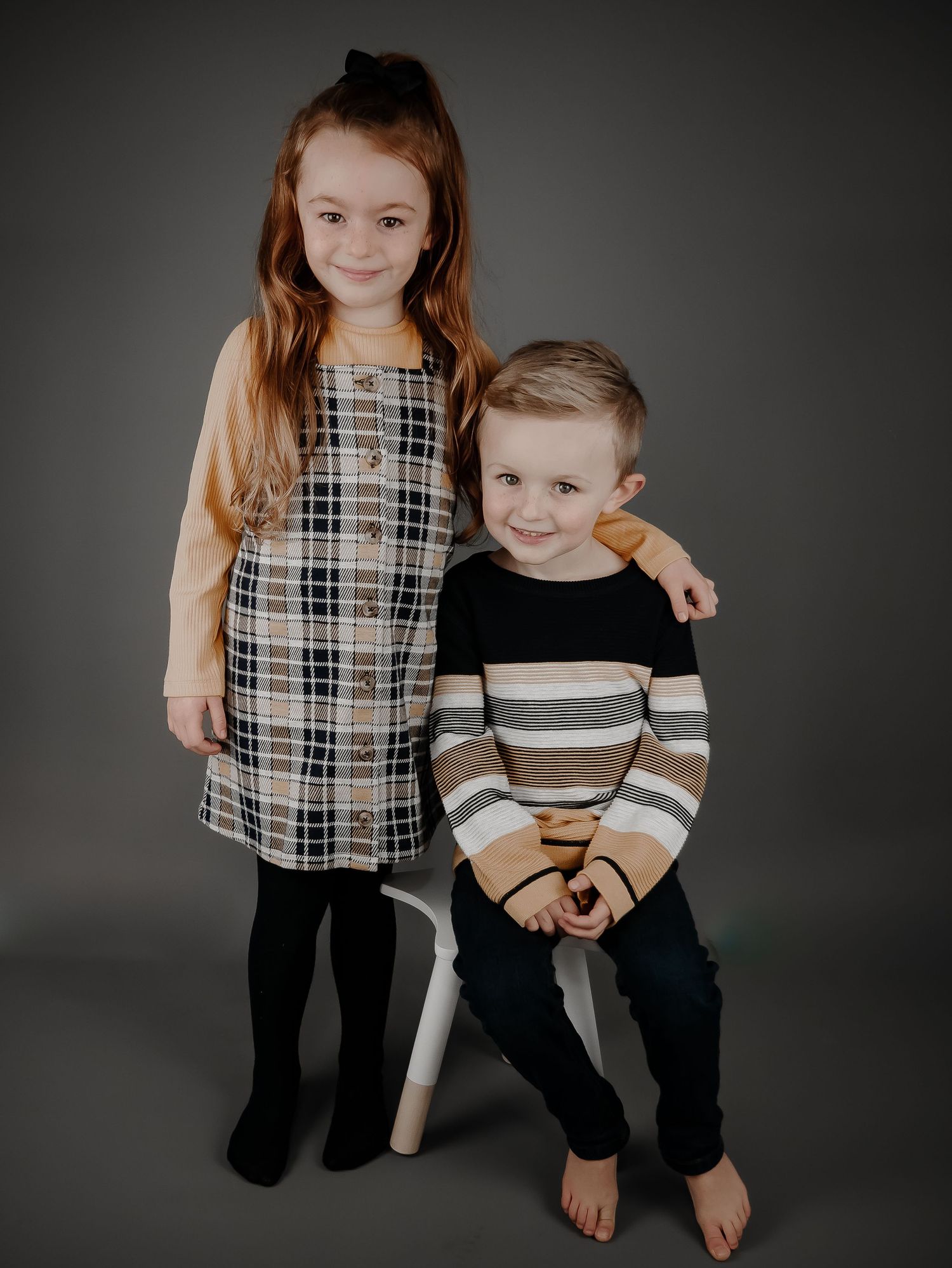 Two children pose together against a gray backdrop, wearing coordinated neutral-toned outfits for a portrait session. sibling photo Cardiff