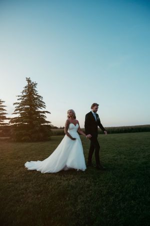 Sunset wedding portrait with couple standing in field near evergreen trees.