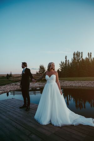 Wedding couple standing on wooden dock by reflecting pool at sunset with trees in background.