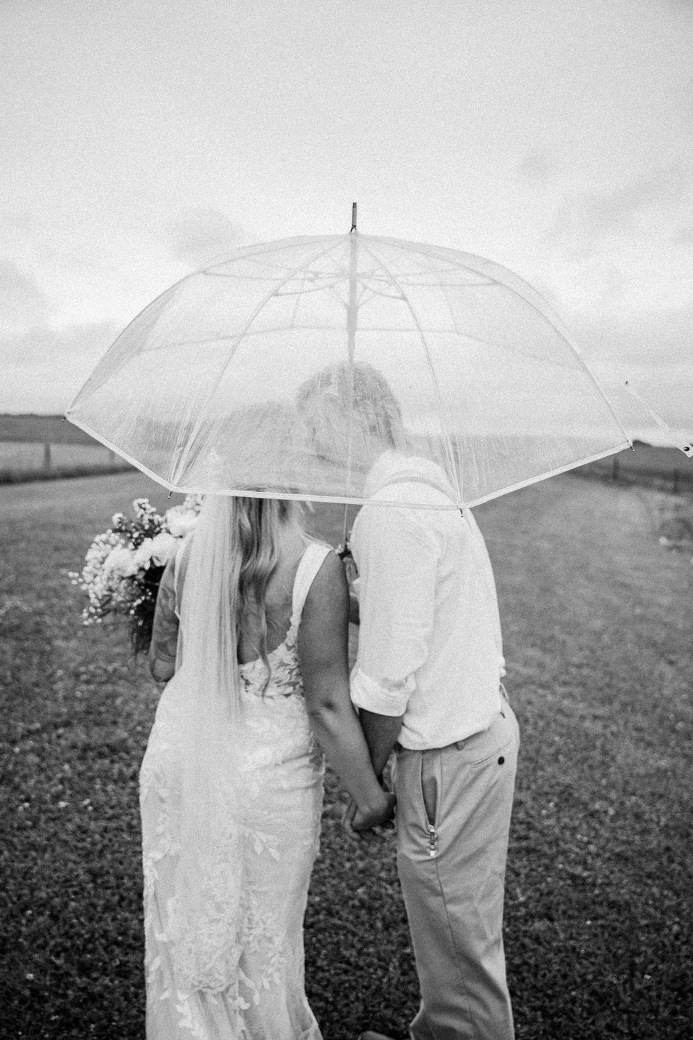 A romantic wedding couple shares a kiss under a clear umbrella in a misty field in black and white.