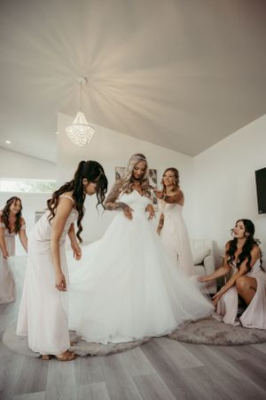 Bridesmaids help with wedding dress preparations in bright white room with crystal chandelier.