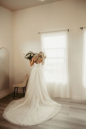 A flowing white wedding dress with a long train photographed in a minimalist room with sheer curtains and natural light.