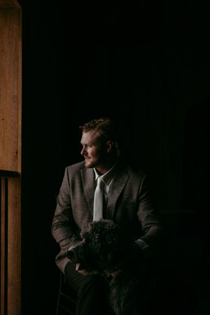 Man in plaid suit holding small dark dog, standing in dramatic low-key lighting near wooden door.