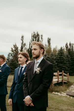 Wedding party members stand in formal attire against backdrop of evergreen trees.