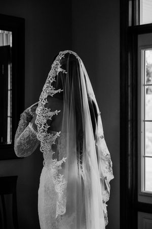Black and white photo of a bride in lace wedding dress and veil standing by window.