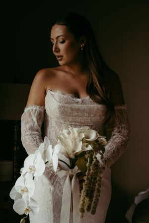 Bride in lace off-shoulder gown holding white orchid and calla lily bouquet with ribbon, posing in soft moody light.