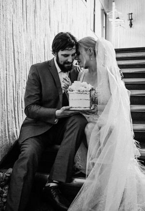 Black and white photo of couple sharing wedding cake on rustic staircase.