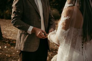 Bride and groom holding hands during outdoor wedding ceremony, bride in lace dress with veil, groom in tweed suit.