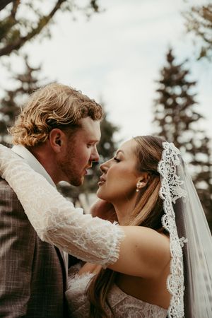 A bride and groom share a romantic almost-kiss moment outdoors, with the bride wearing a lace veil and the groom in a gray suit.