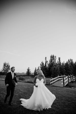 Newlyweds walking on grass path with rustic wooden bridge in background.