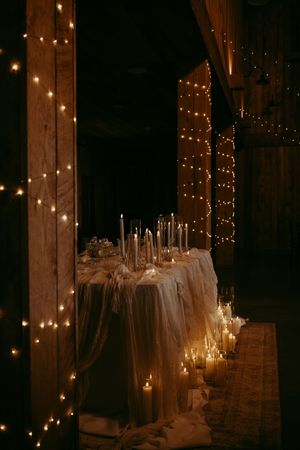 String lights illuminate an elegant wedding reception table setting with white linens and glowing candles in a rustic barn venue.