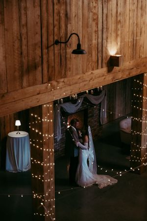 Bride and groom share first dance in a rustic barn venue adorned with warm fairy lights wrapped around wooden beams.