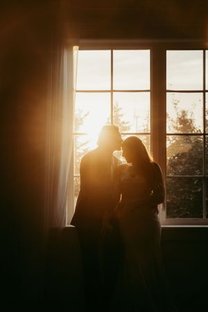 Silhouette of a romantic couple standing together by a sunlit window with warm golden light glowing behind them.