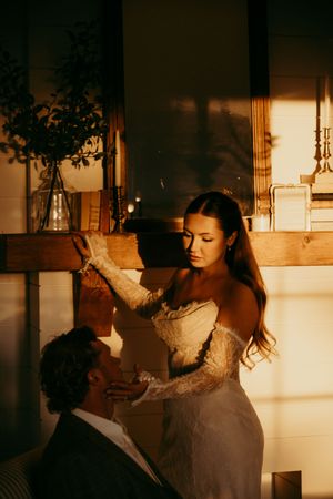 Bride in lace off-shoulder gown tenderly touches groom's face in warm golden light near rustic wooden shelf.