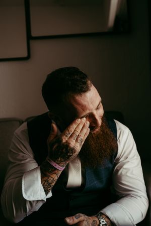 Tattooed bearded groom in white shirt and navy vest emotionally covering his face with tattooed hand on wedding day.
