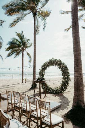 Beach wedding setup with floral hoop arch, Chiavari chairs, and palm trees by the ocean.