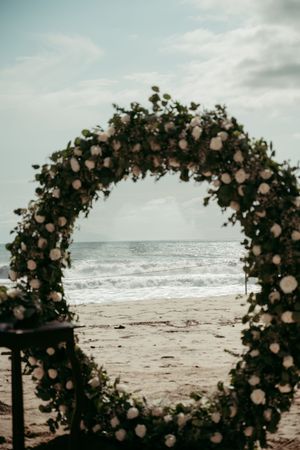 A floral arch with blush roses frames a moody beach view with rolling waves and cloudy skies.
