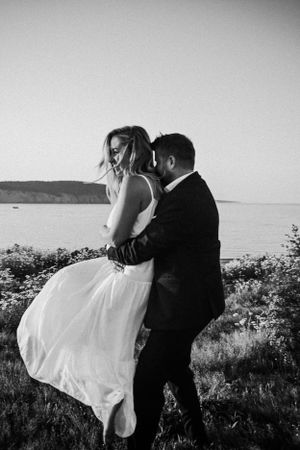 Romantic black and white wedding photo of couple embracing on coastal clifftop with ocean view in background.