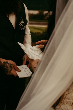 Tattooed groom holds wedding vows card while bride reads handwritten vows during intimate outdoor ceremony.