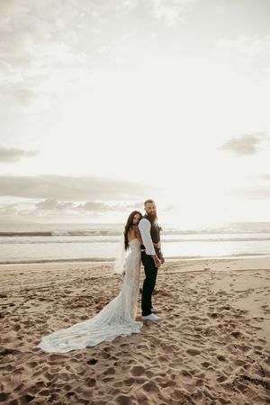 Bride in lace gown and groom holding hands on sandy beach at sunset with ocean waves behind them.