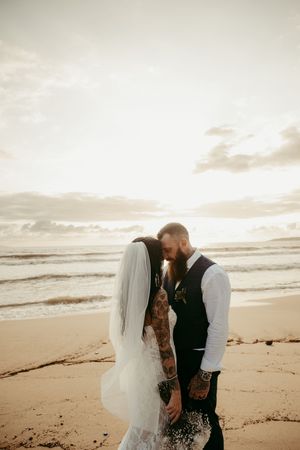 Tattooed bride and groom share intimate moment on sandy beach at sunset, with ocean waves and dramatic sky behind them.