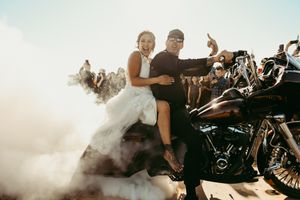 Newlyweds posing on motorcycle with smoke effect and crowd in background.