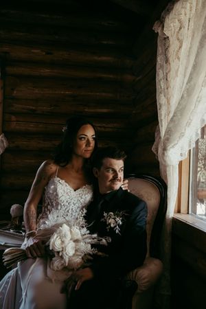 Elegant couple in formal wedding attire sits by window in rustic log cabin with white bouquet.