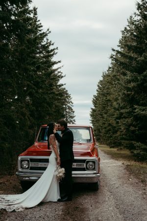 Newlyweds embrace in front of classic red truck on forest dirt road surrounded by pine trees.