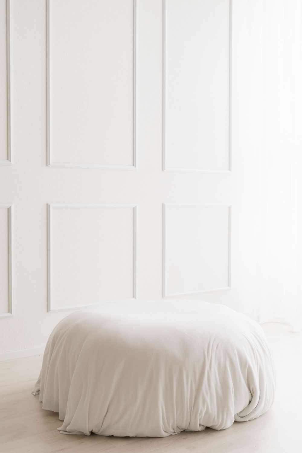 Minimalist white room with a round pouf covered in a cream fabric near a bright window.