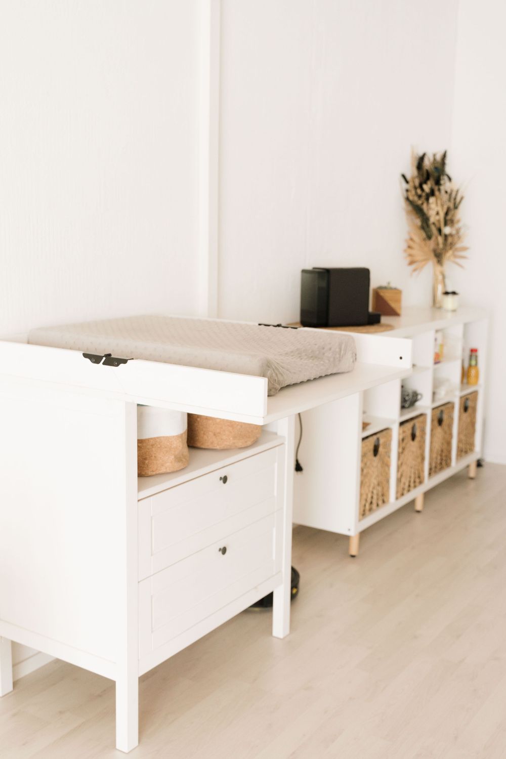 White nursery changing table with wicker baskets and cube storage shelving unit in a minimalist bright room.