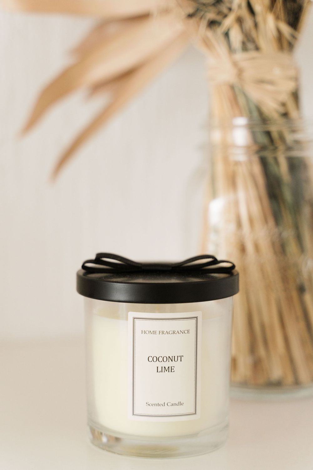 Coconut lime scented candle in glass jar with black bow lid beside dried botanical arrangement.