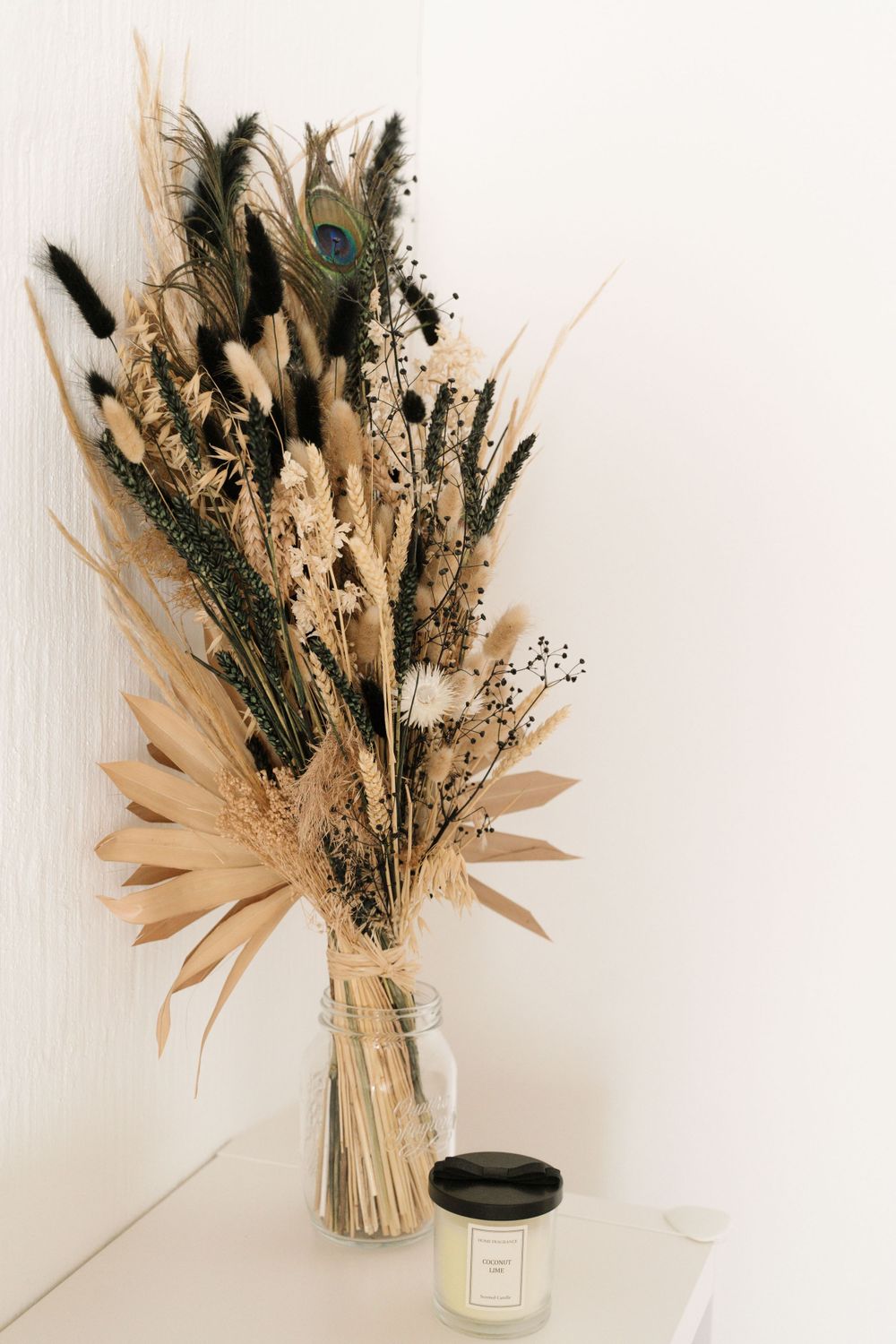Dried floral arrangement with peacock feathers in a glass jar beside a scented candle on a white surface.