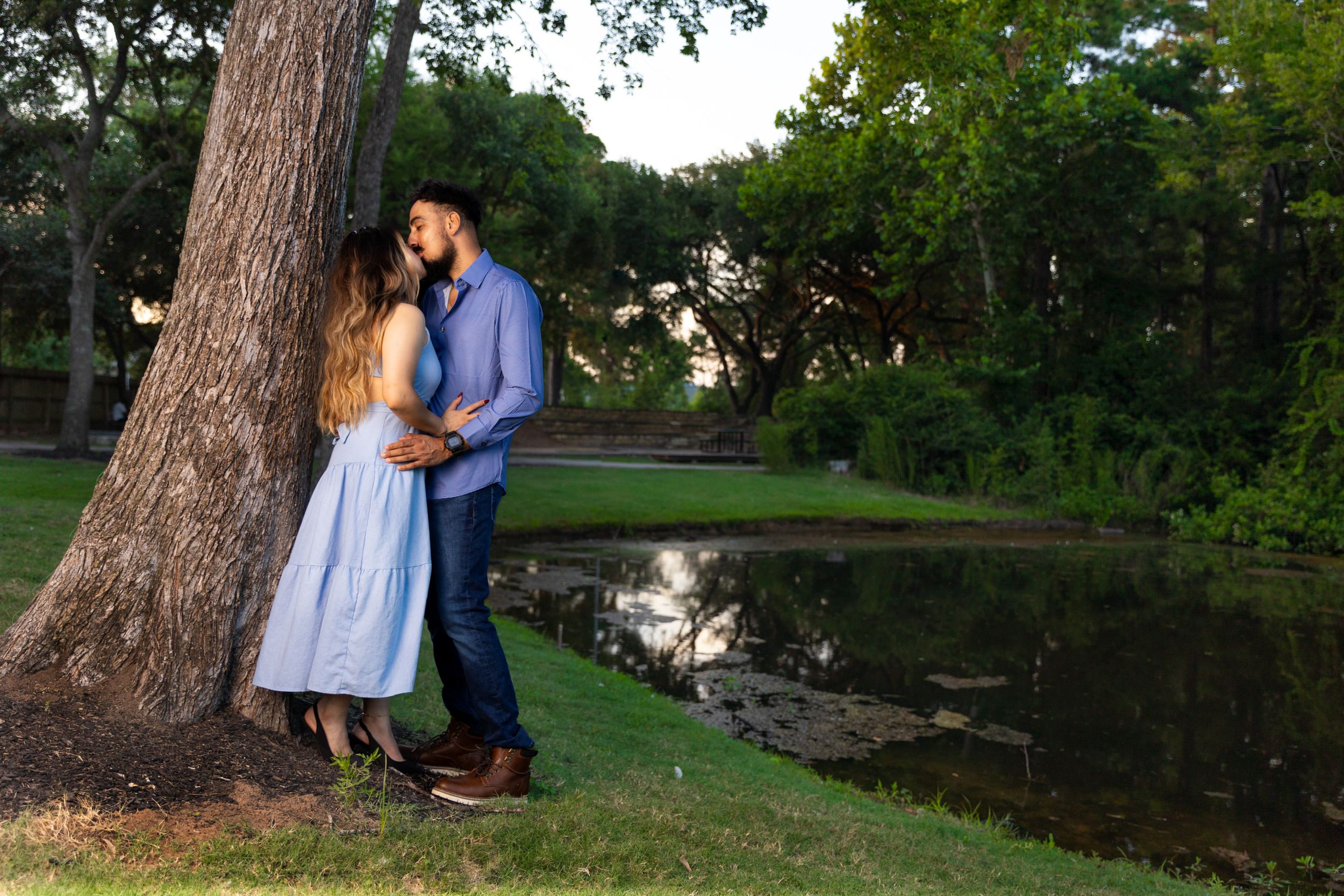 Romantic couple embraces by a tree near a pond during sunset.