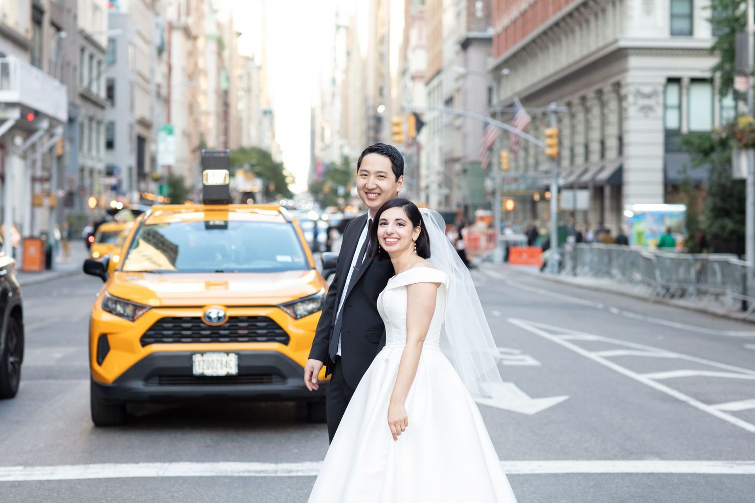 Newlyweds pose on city street with yellow taxi in background.