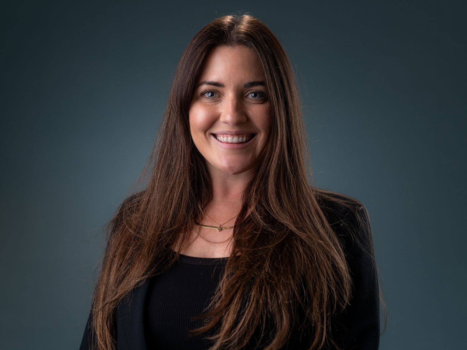 Professional headshot of a smiling woman with long brown hair wearing a black blazer against a dark background.