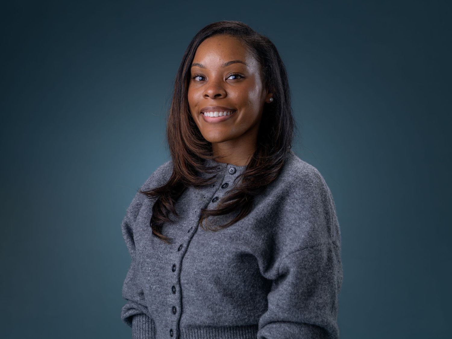 Smiling professional woman with long dark hair wearing a gray cardigan, posed against a dark blue background.