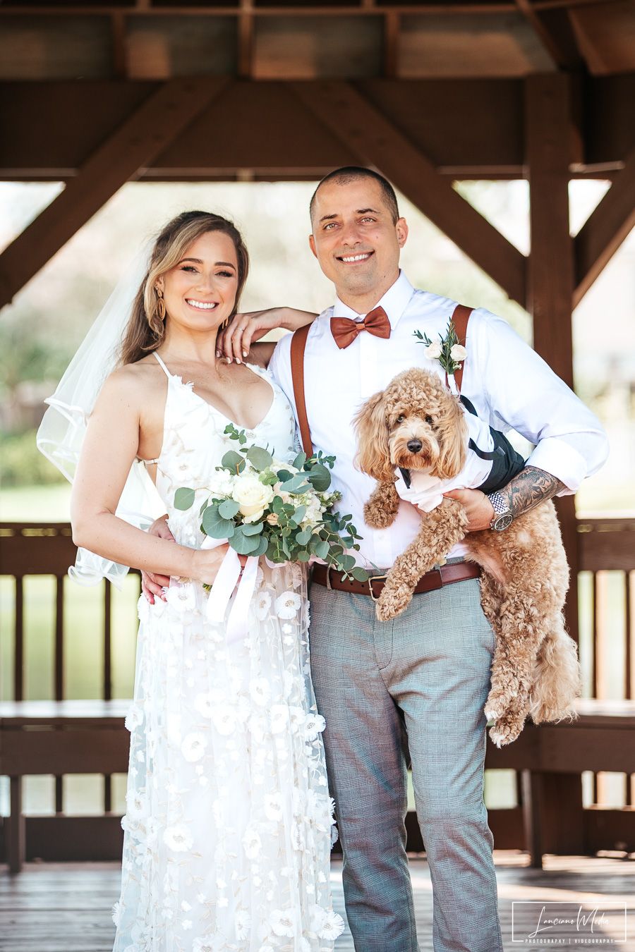 Newlyweds pose with their goldendoodle dog under a rustic wooden pavilion.
