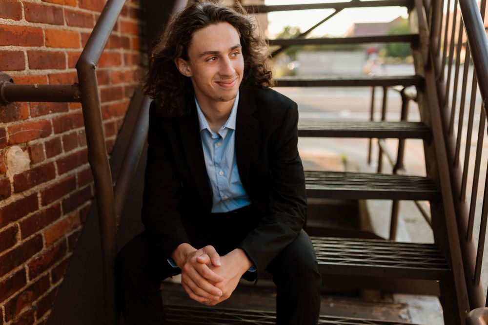 Person in black suit jacket sits on metal stairs near brick wall.