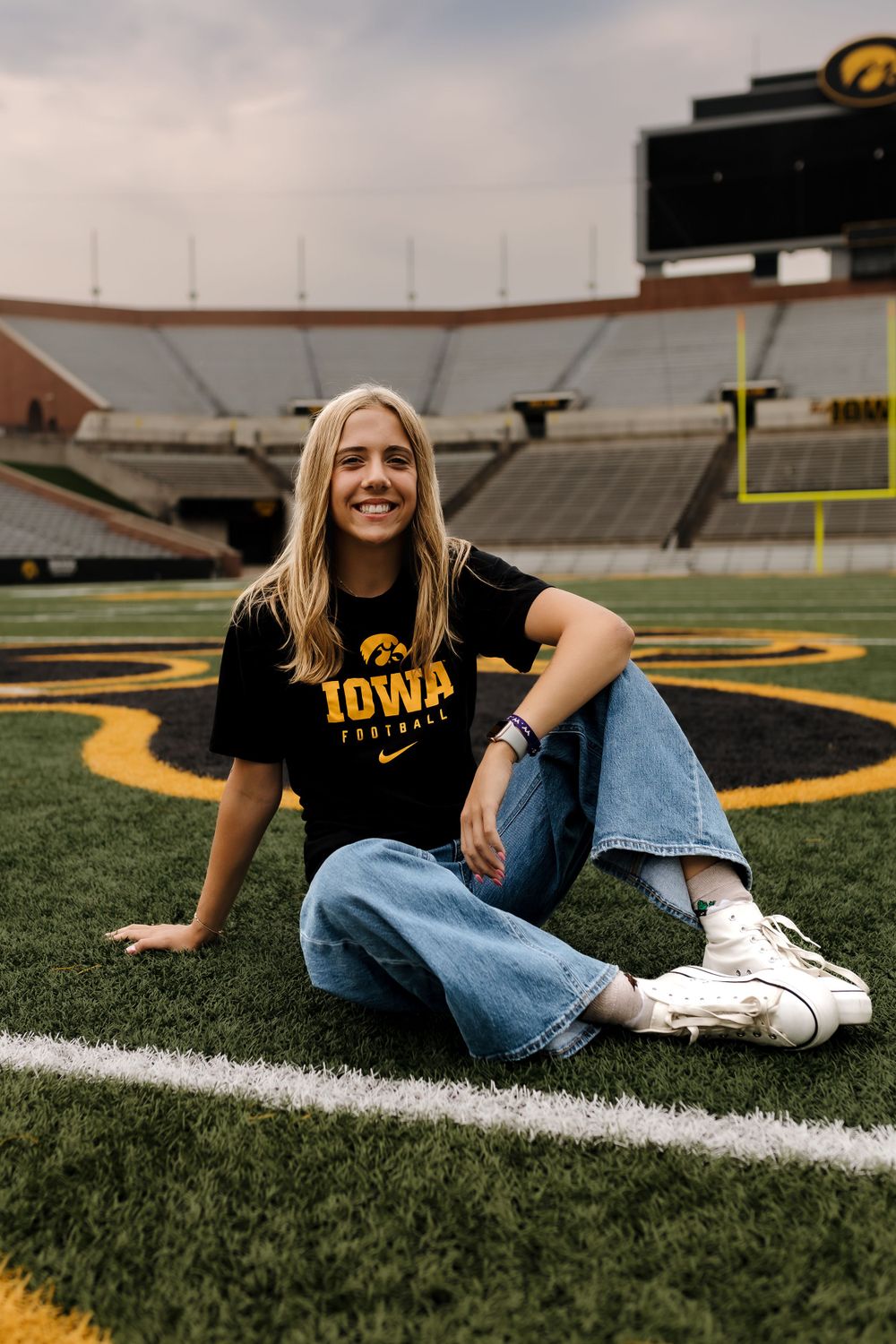 Student in Iowa Hawkeyes shirt sitting on football field with stadium in background.