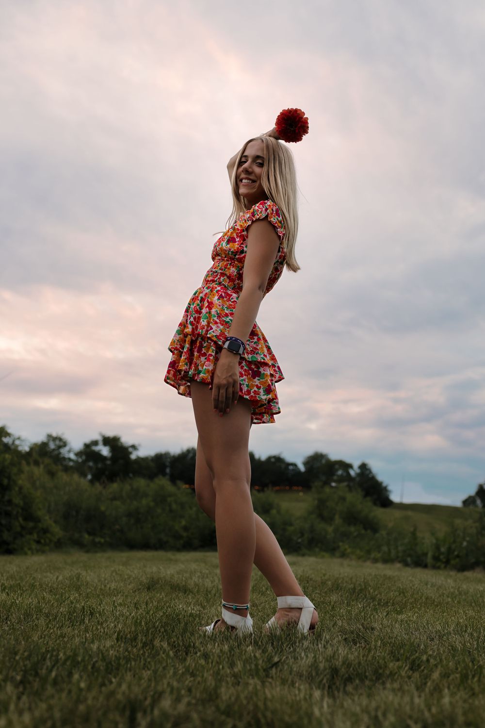 Person in a floral dress poses on a grassy hillside at sunset wearing white sneakers.
