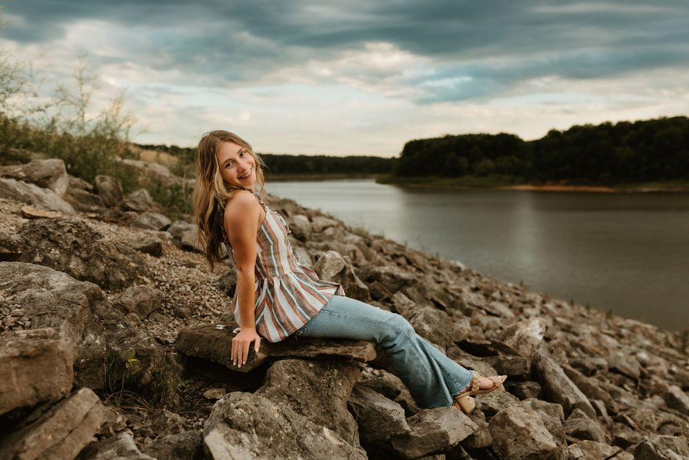 Person in casual attire sitting on rocky shoreline at sunset.