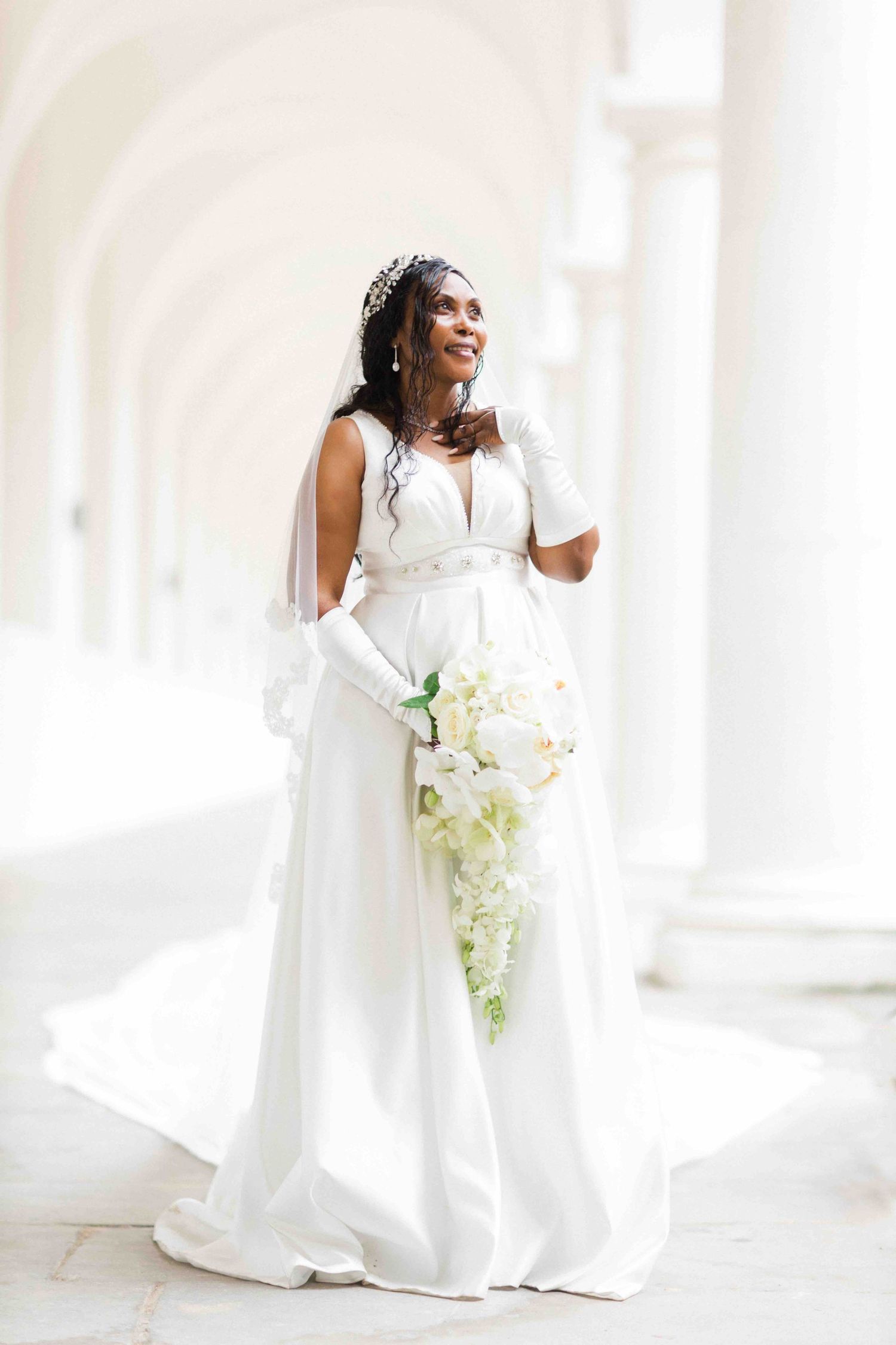 A beautiful bride in a classic white wedding gown holds a cascading bouquet of white orchids in an elegant archway.