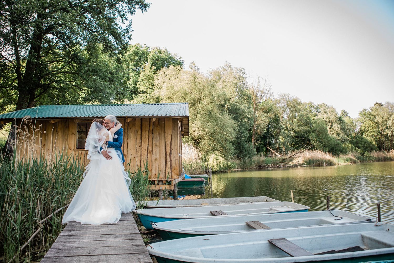 A wedding couple shares a romantic moment on a wooden dock with boats and a rustic cabin by a serene lake.