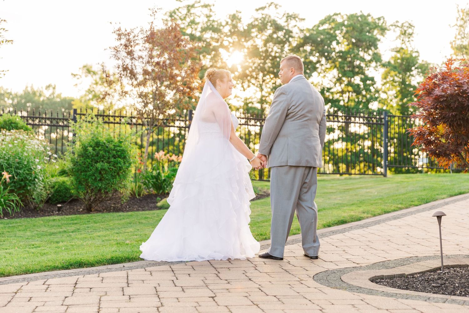Couple in wedding attire walking along paved garden path at sunset.