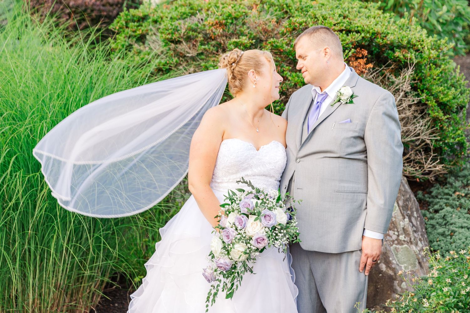 A couple in wedding attire stands near lush greenery with the bride's veil flowing in the breeze.
