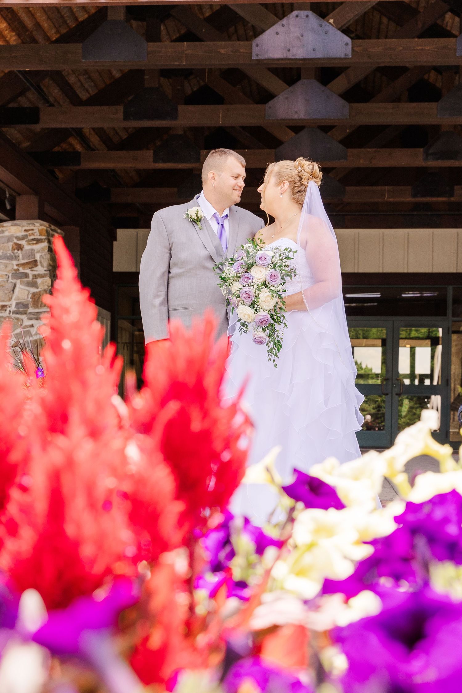 A wedding scene framed by vibrant red and purple flowers under a wooden pavilion.
