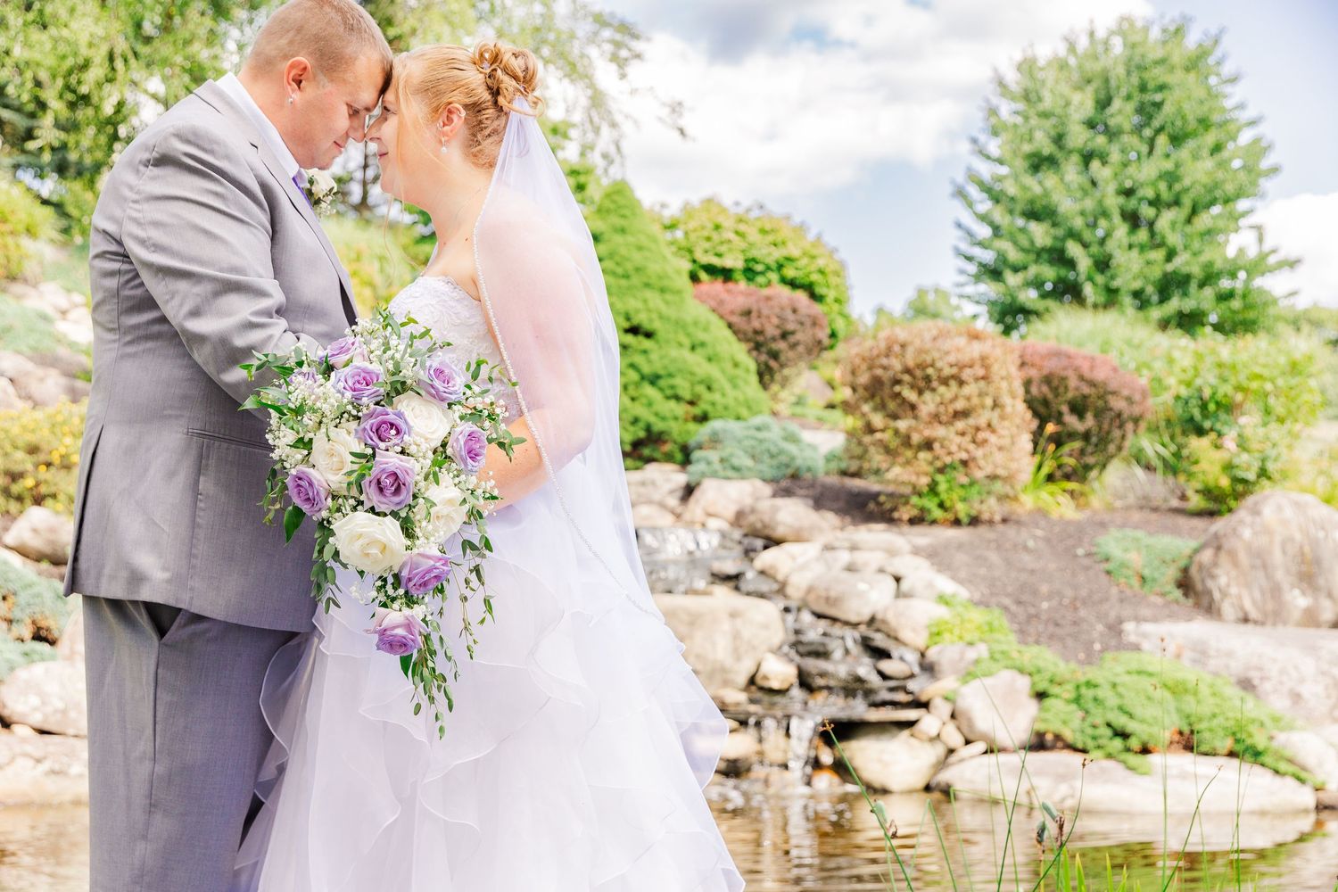 Couple poses for portrait by decorative water feature with purple floral bouquet.